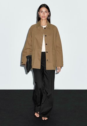 Brown cotton jacket with large buttons, paired with a light top and loose black pants. A textured black clutch is held in one hand.