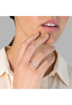 Anello d'argento con un centro di pietra preziosa trasparente e rotonda, indossato su una mano che tocca delicatamente il mento. Banda liscia con una texture sottile.