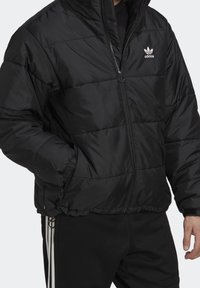 Czarna kurtka puchowa zapinana na zamek z przodu, z ściągaczami na mankietach i regulowanym dołem. Na piersi znajduje się logo Adidas, wykonana z gładkiego, błyszczącego materiału.