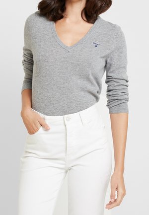 Frau trägt einen hellgrauen V-Ausschnitt-Pullover mit GANT-Logo und hoch taillierte weiße Hose, eine Hand in der Tasche, vor einfachem hellem Hintergrund.