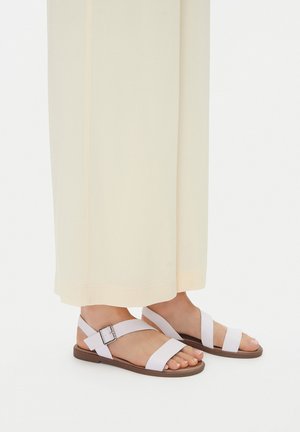 Pies con sandalias planas de cuero blanco, con dos tiras anchas y una hebilla, combinadas con pantalones anchos color crema.