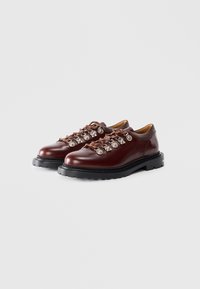 Burgundy nahkakengät, joissa on metalliset silmukat, ruskeat nauhat ja paksu musta kumipohja, jossa on tekstuurinen kuvio.