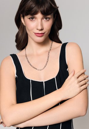 Czarna i biała w paski bezrękawnik z okrągłym dekoltem. Modelka nosi srebrny naszyjnik łańcuszkowy. Ręce w odcieniu skóry skrzyżowane na talii.