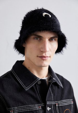 Fekete bolyhos vödörkalap fehér ívelt díszítéssel; fekete farmerdzsekivel viselve, amelyen kontrasztos fehér varrások és gombos záródás található.