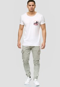 Camiseta blanca de manga corta con un gráfico de una palmera en el bolsillo del pecho izquierdo, combinada con pantalones cargo verde claro y zapatillas blancas.