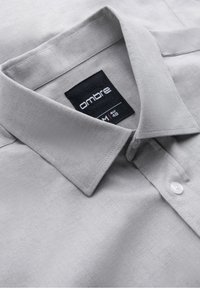 Chemise habillée gris clair avec col pointu, bouton supérieur visible et étiquette de marque noire portant la mention "ombre" taille M EUR 48.