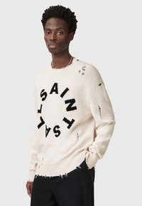 Cremefarbener Pullover mit schwarzem "SAINT"-Text in kreisförmigem Muster. Verfügt über ausgefranste Details, einen runden Halsausschnitt und gerippte Bündchen.