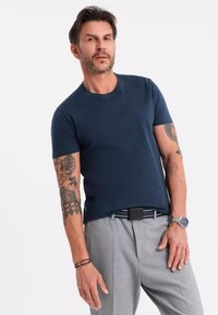 T-shirt di cotone blu navy con maniche corte, colletto a giro, indossato con pantaloni grigi chiari su misura e una cintura a strisce, che mette in mostra tatuaggi visibili.