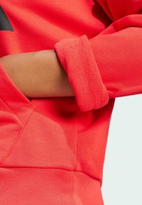 Röd sweatshirt med en strukturerad tyg, ribbade ärmslut och en känguruficka, som visar en modern design med en ren silhuett.