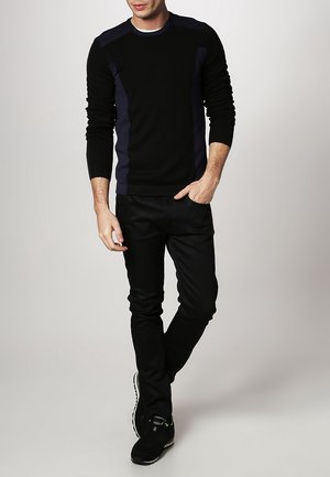 Sort sweater med mørkeblå sidepaneler, rund halsudskæring og ribstrikkede manchetter, kombineret med sort slim-fit jeans og sorte sportslige sko.