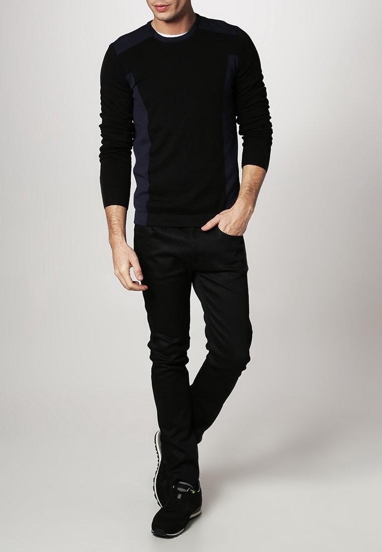 Pull noir avec des panneaux latéraux bleu foncé, col rond et poignets côtelés, associé à un jean noir slim et des chaussures de sport noires.