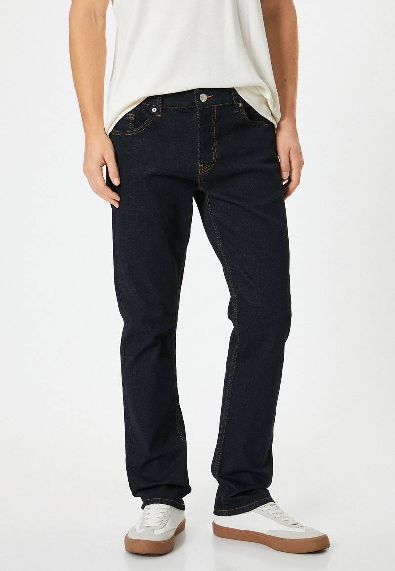 Jeans azul oscuro con costuras naranjas, de corte slim y diseño clásico de cinco bolsillos, combinados con zapatillas blancas y una blusa clara.