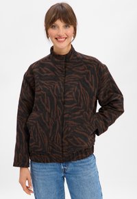 Woman smiling wearing a brown and black tiger-striped jacket with two front pockets and blue jeans against a plain white background.