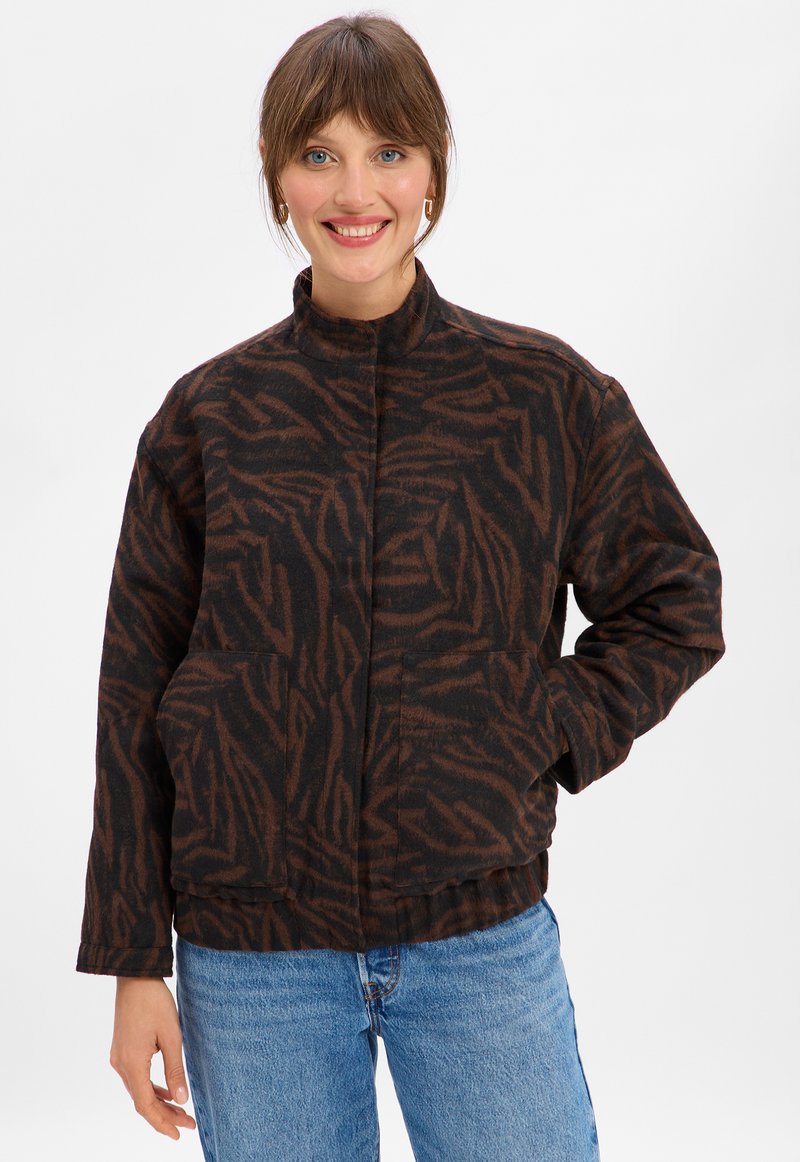 Woman smiling wearing a brown and black tiger-striped jacket with two front pockets and blue jeans against a plain white background.