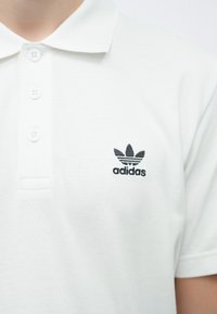 Λευκό πόλο πουκάμισο από υφασμάτινη υφή, με μαύρο λογότυπο Adidas στο αριστερό στήθος και τρία κουμπιά στο γιακά.