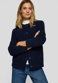 Marineblauer, strukturierter Cardigan mit rundem Ausschnitt, vorderer Knopfleiste und zwei Fronttaschen, kombiniert mit einem weißen Top und Jeans.