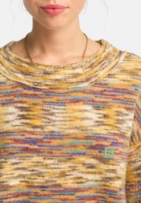 Sweter z dzianiny z wielokolorowym wzorem w odcieniach żółtego, brązowego i niebieskiego. Szeroki dekolt i mała zielona łatka z logo z przodu.