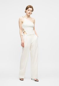 Γυναίκα που φοράει λευκό τοπ με έναν ώμο και λεπτομέρειες από χρυσούς κρίκους, καθώς και λευκό παντελόνι ψηλόμεσο, φαρδύ, με τα χέρια στις τσέπες.