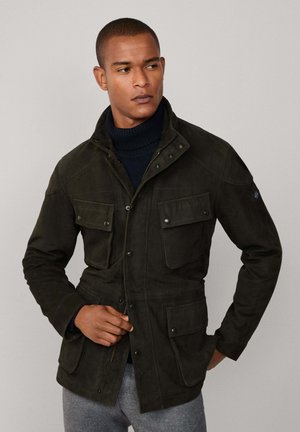 Dunkelgrüne, strukturierte Jacke aus Wildleder mit hohem Kragen, mehreren Fronttaschen und Druckknopfverschlüssen.