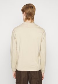 Beige Langarm-T-Shirt aus weichem Baumwollmaterial, mit Rundhalsausschnitt und geradem Saum. Getragen mit dunkelbraunen Hosen.
