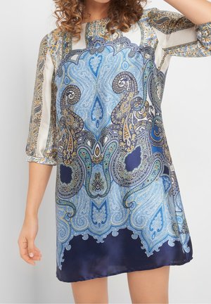 Femme portant une robe ample avec des motifs paisley et floraux complexes en bleu, blanc et or, manches trois-quarts, debout devant un fond blanc.