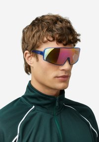 Gafas de sol azules de gran tamaño con lentes espejadas degradadas, con un diseño angular y elegante. El modelo lleva una chaqueta verde oscuro con cremallera.