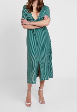Kobieta ubrana w zieloną i białą sukienkę midi w pionowe paski z głębokim dekoltem w serek i rozcięciem z przodu, w połączeniu z białymi sandałami na wysokim obcasie.