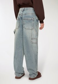 Osoba ubrana w luźne, jasnoniebieskie dżinsy z dużymi tylniami i bocznymi kieszeniami, w połączeniu z brązowymi butami i ciemną górą.