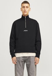 Black zip-up sweatshirt with high collar, ribbed cuffs, and logo on chest, worn over a light-colored shirt and paired with gray jeans.