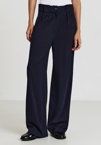 Dunkelblaue, weit geschnittene Hose mit Nadelstreifenmuster, hoher Taille, vorderen Falten und einem einzelnen Knopfverschluss. Glatter Stoff und gerader Schnitt.