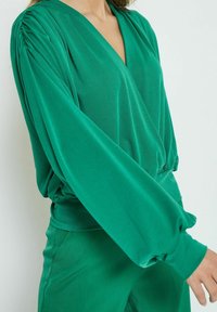 Maglietta verde a maniche lunghe con scollatura a V profonda, dettagli arricciati sulle spalle e vestibilità rilassata, realizzata in un morbido e leggero tessuto.