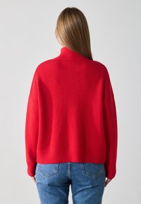 Suéter rojo de punto con cuello alto, textura acanalada y ajuste holgado, combinado con jeans de mezclilla azul, vista trasera.