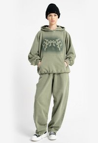 Osoba w luźnej oliwkowej bluzie z kapturem i pasujących spodniach, w czarnej czapce beanie i białych sneakersach, stojąca z rękami w kieszeniach.
