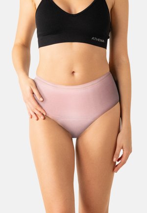 Femme portant un soutien-gorge de sport noir et une culotte taille haute rose poudré, devant un fond clair uni.