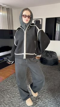 Črna jopa z zadrgo z belimi poudarki, črne trenirke in svetlo rjavi copati. Model nosi sivi šal in temna sončna očala. Pod njim je puhast sivi preproga.