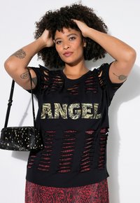 Černé tričko s výřezovým designem, nápis "ANGEL" v leopardím potisku, červené detaily. Modelka drží malou černou kabelku s tečkovaným vzorem.