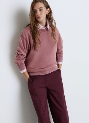 Donna con capelli lunghi e ricci che indossa una felpa rosa sopra una camicia rosa chiaro con colletto e pantaloni bordeaux con le mani in tasca.