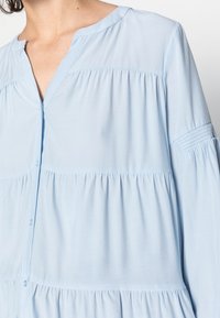 Camisa azul clara, de botão, com um corte relaxado, apresentando seções franzidas e um decote em V. Feita de tecido leve e macio.