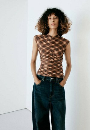 Femme aux cheveux bouclés portant un top sans manches marron à motifs froncé et un jean bleu foncé ample, les mains dans les poches.