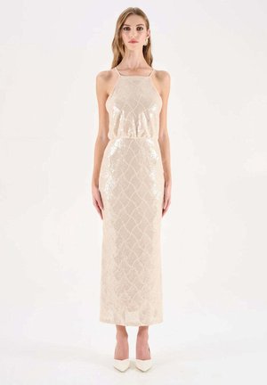 Donna che indossa un abito senza maniche, lungo fino al pavimento, color oro pallido con paillettes, collo alto e tacchi bianchi appuntiti su sfondo bianco.