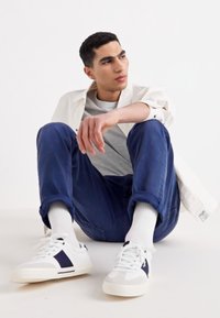 Conjunto casual que incluye una camiseta gris, una chaqueta blanca, pantalones azul marino y zapatillas blancas con detalles en azul marino. Modelo sentado sobre un fondo claro.