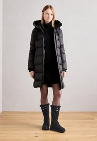 Abrigo acolchado negro con capucha, diseño acolchado y longitud larga, combinado con botas negras con cordones que tienen un borde de piel de peluche y suelas de goma.