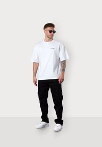 Weißes Baumwoll-T-Shirt in Übergröße mit Logo, kombiniert mit schwarzen Cargo-Hosen. Die Accessoires umfassen schwarze Sonnenbrillen und weiße Sneakers.