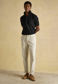 Un homme se tient debout, les mains derrière le dos, portant un polo noir à manches courtes, un pantalon beige et des chaussures marron à lacets, devant un fond beige.