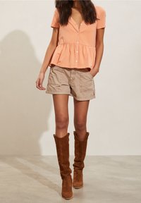 Blusa pesca a maniche corte con bottoni sul davanti e vita a peplo, abbinata a pantaloni cargo beige e stivali in suede marroni fino al ginocchio.