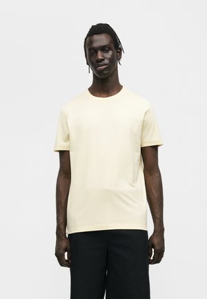 Mand med mørk hud og fletninger iført en ensfarvet kortærmet creme t-shirt og sorte bukser, der står mod en lys baggrund.