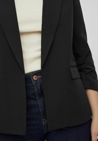 Černý blazer se strukturovanými klopami a detailem jednoho knoflíku, vrstvený přes krémový top a tmavé džíny s měděným knoflíkem.