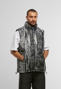 Gilet à motifs noir et blanc avec col montant, fermeture éclair et poches latérales ; présente des designs géométriques et abstraits intriqués.