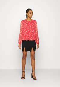 Top floral vermelho com mangas longas transparentes, combinado com uma saia preta justa com uma fenda frontal. Usado com sandálias de salto pretas.