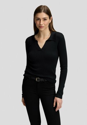 Kvinde med glat brunt hår iført en tætsiddende sort V-hals ribbet sweater, sorte bukser og et sort bælte, stående mod en ensfarvet baggrund.